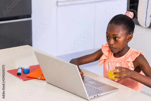 African American child working at kitchen counter using laptop holding orange juice, copy space