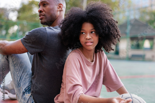 Father and daughter sit back-to-back with a basketball.