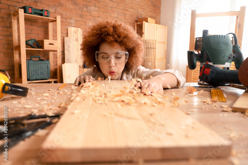 Bild auf Leinwand Young female carpenter blowing wood shavings from plank in workshop