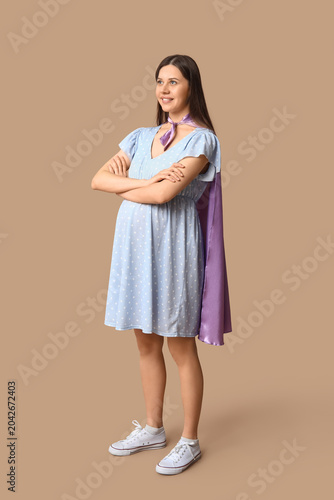 Beautiful young pregnant woman in superhero cape on brown background