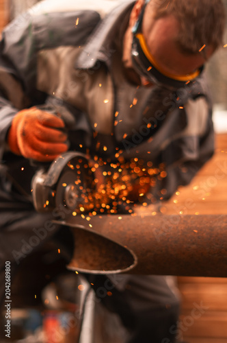 Construction specialist cuts damaged steel pipeline with angle grinder during repair operation outdoors. Bright sparks, industrial tools illustrate infrastructure maintenance and engineering service