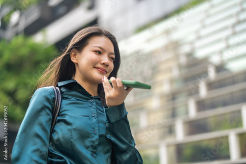 Smiling asian business woman using AI voice assistant on smartphone outdoors, Smart agent technology, Female corporate employee recording audio message or speaking to artificial intelligence app