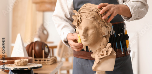 Fotografi Young male sculptor with sponge shaping clay bust in workshop, closeup
