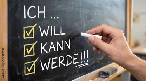 A hand writes motivational german words on a classroom chalkboard with white chalk to represent the progression from wanting to being able and finally achieving