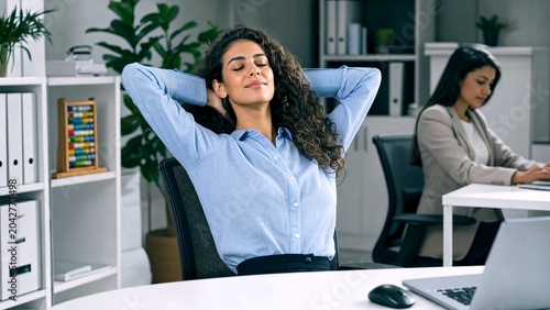Relaxed businesswoman leaning back in office chair with hands behind head, calm female employee taking a break at work desk, peaceful corporate workspace