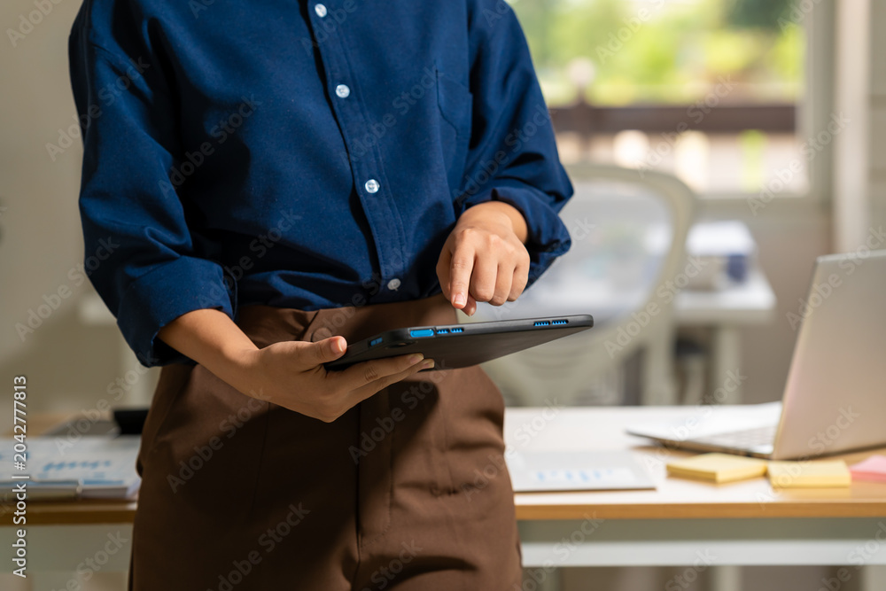 Fototapeta premium Business professional standing in a bright office and using a tablet near a desk with laptop and notes, showing modern work, planning, communication and flexible office life.