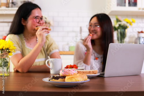 Close-up view of delicious sweets and donuts on a work desk, with the background showing two best friends happily enjoying donuts and strawberries while working on their laptops in the kitchen at home