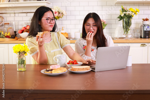 Two happy young Asian women enjoying snacks and desserts while working on a laptop in a cozy kitchen. Friendship, lifestyle, and remote work concept.