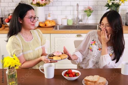 Young woman refusing donuts for a healthy diet and weight loss. She shows a hand gesture to stop while her friend offers sweets in the kitchen. Concept of healthy lifestyle.