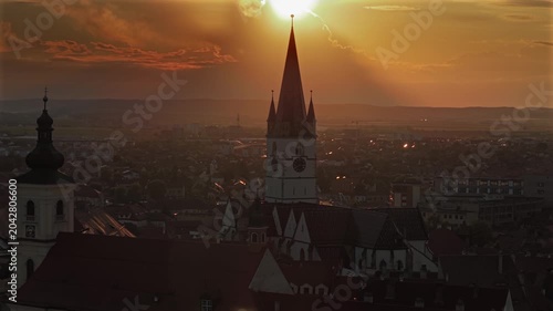 Aerial 4k video of the old town of Sibiu featuring historic red roofs and the tall church tower, with the majestic blue Fagaras mountains visible on the horizon. Sibiu, Transylvania, sunset landscape.