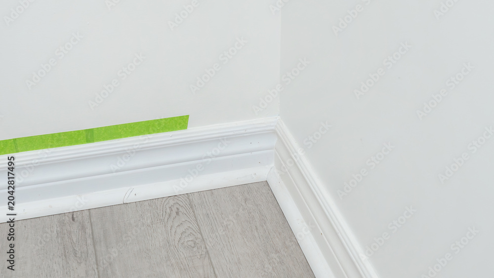 Fototapeta premium Corner of a room with freshly painted white walls, wooden floor, and green painter's tape applied along the baseboard for clean edges