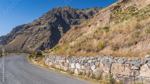 The asphalted highway bends around the mountain. There is dry yellow grass on the slope. A stone barrier along the road. The blue sky. Peru.