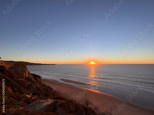 Schilderij op canvas Ocean sunset with sun reflecting on water and rocky headland silhouette