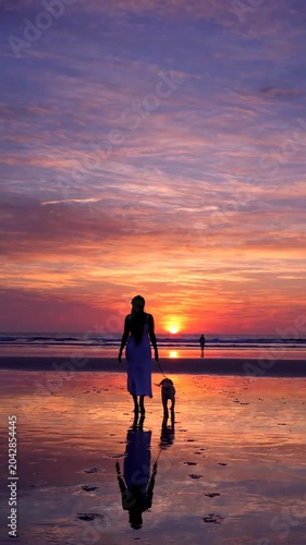 Woman walking on beach with dog at sunset.