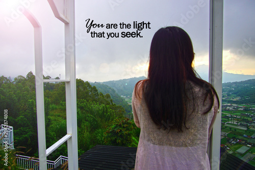 Inspirational motivational quote - You are the light that you seek. With young woman standing alone from behind looking at the mountain view in the morning at sunset. Self love care and confidence.