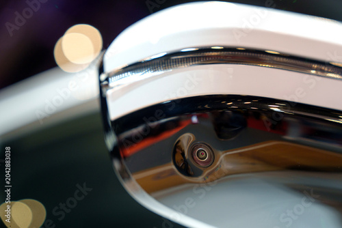 A detailed close-up shot of an surround-view system camera built into the lower black gloss housing of a white side wing mirror on a vehicle displayed at an automotive exhibition.