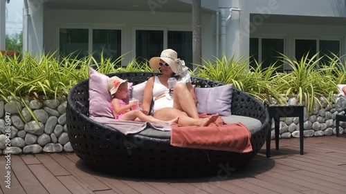 Mother in straw hat and white swimsuit relaxing with her little daughter in pink swimsuit on a poolside lounge chair drinking fruit cocktails. Family holiday concept