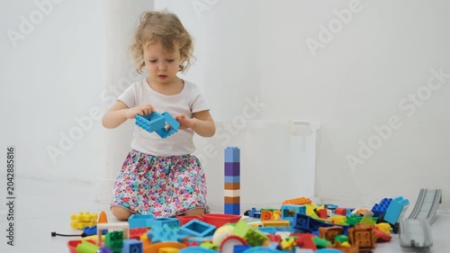 Cute little girl in a floral skirt sitting alone carefully connecting colorful plastic block pieces together on a play floor. Childhood and development concept