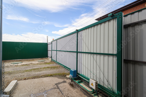 long sliding metal gate with a green frame and silver panels, equipped with a blue electric drive and control box, set against a corrugated fence on a concrete driveway