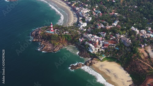 Aerial view of the iconic red and white Vizhinjam Lighthouse on a rocky headland overlooking the sandy beaches and turquoise waters of Kovalam, Kerala, India.