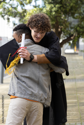 Diverse males hugging on path, graduate in gown holding diploma and cap, friend in denim jacket