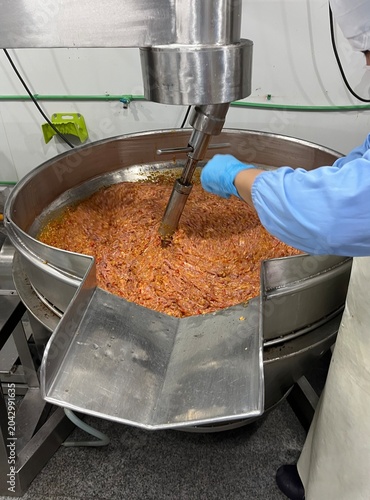 Worker operating a large industrial mixing kettle during food manufacturing. Food ingredients being mixed and processed to ensure product consistency and quality in large-scale food production.