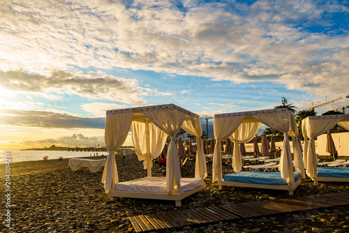Tableau sur toile White beach cabanas on pebble shore under dramatic cloudy sky