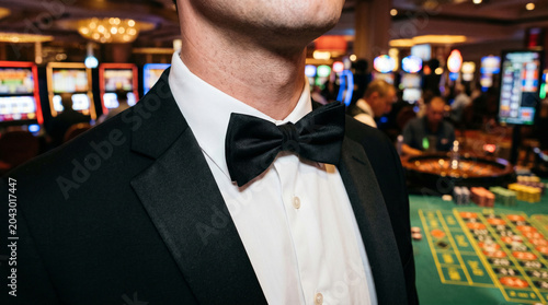Close-up of a casino employee wearing a formal tuxedo and bow tie with strongly blurred slot machines and a roulette table in the background, representing gaming, luxury nightlife, and entertainment