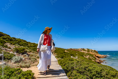 Middle-aged woman walking on beach boardwalk over ocean on sunny summer day. Front view