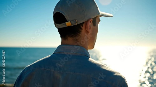 Young man in a blue cap and shirt looking at the calm ocean under a bright clear sky