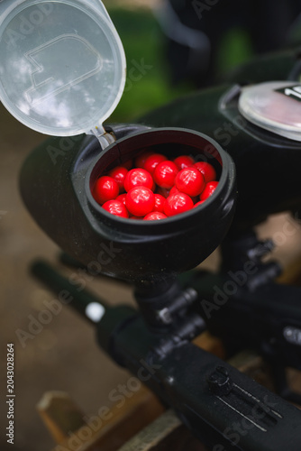 Close-up of paintball marker hopper filled with red paintballs before outdoor game. Recreational shooting equipment prepared for team competition and tactical sport activities.