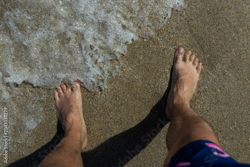 Two feet of a white man on the wet sand of a beach as ocean waters come near by.
