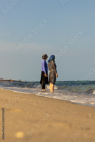 Two fully dressed women in dresses and head scarfs standing on the beach as ocean water waves hit their feet.