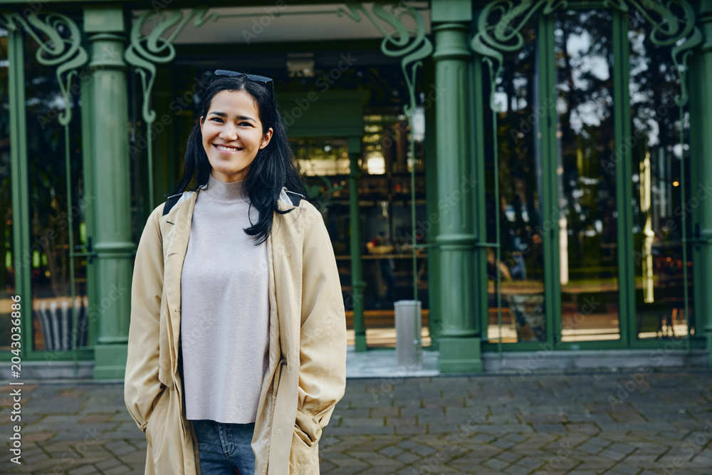 Naklejka premium Woman in beige coat and gray sweater stands outdoors in front of a green storefront, smiling in an urban daylight scene, blending casual fashion with a relaxed city mood and bright daytime ambiance.