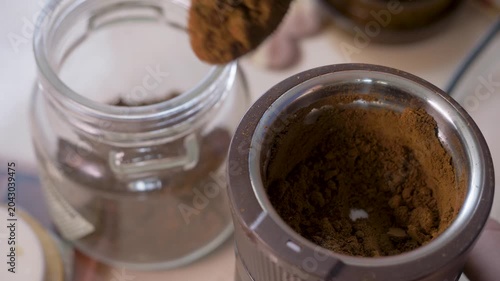 Hand fills container with fresh coffee powder from grinder. Glass jar holds aroma meant to energize mornings and improve focus. Roasted coffee beans have been ground down to be brewed for coffee