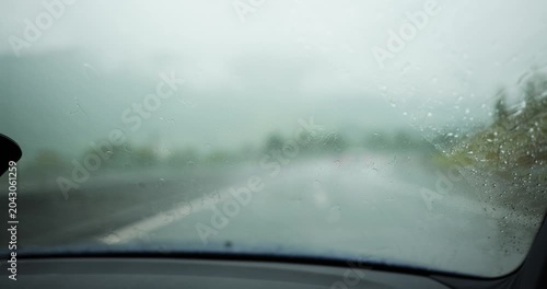 Interior point of view shot looking through a car windshield during a heavy downpour, showing windshield wipers actively clearing rainwater while driving on a blurred highway road