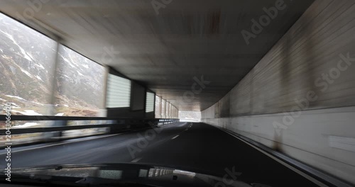 Point of view shot from inside a car driving through a concrete semi-tunnel or avalanche protection gallery on a winding mountain road, with a scenic view of snowy alpine slopes