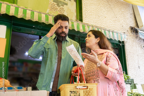 Young Indian couple shocked reading grocery receipt or bill due to high cost of goods and rising inflation problem in india at supermarket.