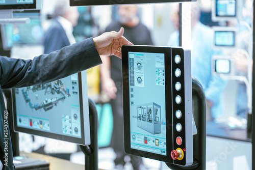 A person points toward an industrial human-machine interface (HMI) monitor displaying manufacturing metrics and diagnostic diagnostics at an automation trade fair.