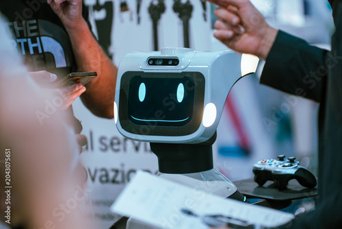 An interactive service robot with a digital smiley face display is shown during a product demonstration at an industrial automation trade fair, as visitors and representatives interact nearby.