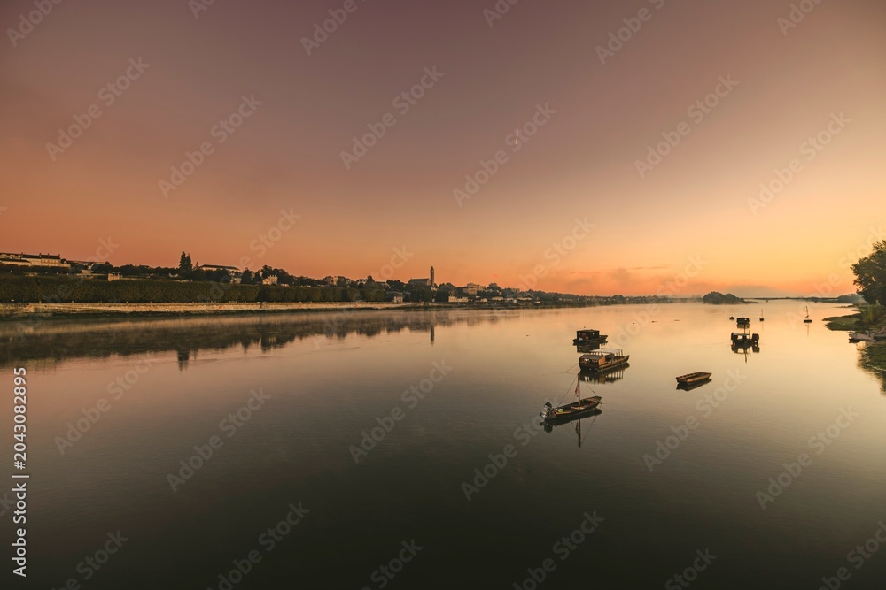 Fototapeta premium As the sun begins to set, serene waters mirror the sky's vibrant hues. Small boats drift gracefully on the river, with hills framing a picturesque scene of peace and beauty