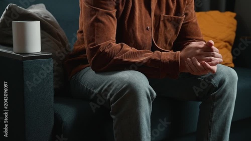 A man in casual clothing sits on a couch with his hands clasped together. He appears pensive as he reflects in a well-lit living space with soft furnishings around him