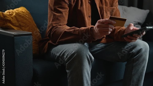 A person sits on a couch while holding a credit card and a remote in a living room. The individual is engaged in an online shopping activity, indicating a modern lifestyle