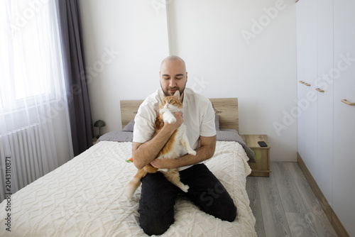 Schilderij op canvas Young adult man sitting on a sofa and caresses ginger tabby cat