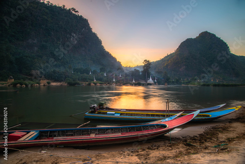 Fotografi A pier with fishing boats along the river, situated in a natural setting with a pagoda and mountains