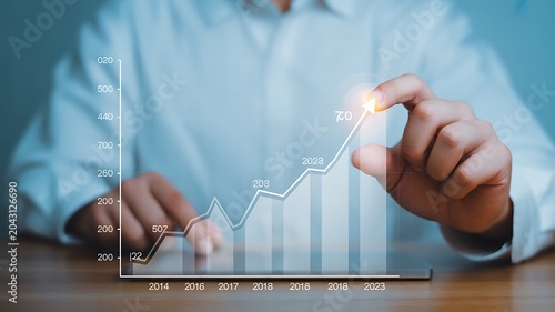 Business growth chart showing upward trend, with hand pointing to the peak on a digital screen illustrating financial success and positive development over time
