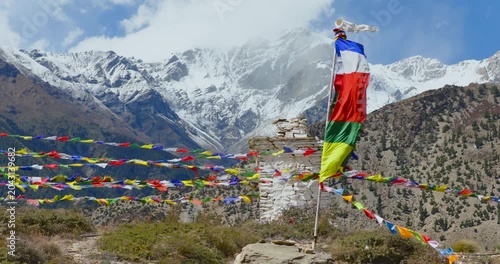 Sideways drone shot capturing a traditional Buddhist chorten with vibrant Lung ta and Darchog flags in Mustang, Nepal, as it is fluttering intensely against a cold, misty Himalayan mountain