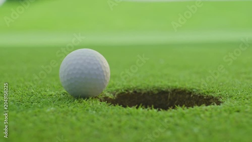Macro 4K video of a white golf ball rolling on a lush green putting green and stopping right on the edge of the hole, showcasing a dramatic near miss, suspenseful game, and sports concept.