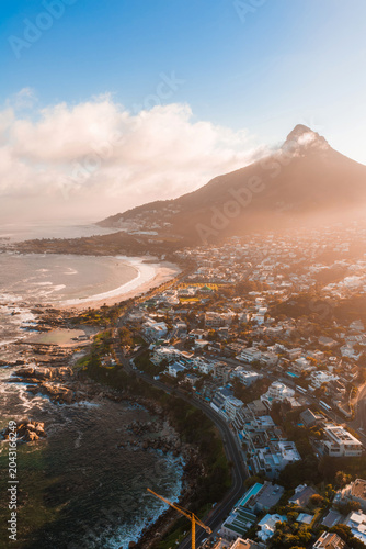 Aerial view of Cape Town's Atlantic Seaboard shows Lion's Head, Camps Bay, and Clifton at sunset. Warm light bathes white sand and rocky outcrops as low clouds drift.