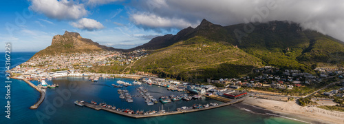 Tableau sur toile Aerial view of Hout Bay Harbour in Cape Town shows marina, fishing boats, curved breakwater, sandy beach, and the Sentinel under broken clouds and Table Mountain slopes